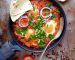 Shakshuka with pita bread in a pan. Middle eastern traditional dish. Fried eggs with tomatoes, bell pepper, vegetables and herbs. Shakshouka on a table. Top view. Sunny side up eggs. From above